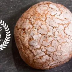 Chrupiący bochenek chleba orkiszowego z popękaną skórką, obok wieniec laurowy z napisem "Przepis na chleb orkiszowy".