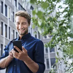 Uśmiechnięty mężczyzna w granatowej koszuli korzysta z telefonu na tle budynku i zieleni.