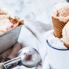 Due palline di gelato fatto in casa, con pezzetti di fragola, in un cono, accanto a una vaschetta e un porzionatore.