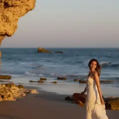 Uśmiechnięta kobieta w białej sukience, trzymająca kapelusz, spaceruje po plaży. Idealny przykład, jak pozować do zdjęć nad morzem.