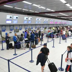 Długa kolejka ludzi z bagażami czeka na odprawę na lotnisku. W tle widać stanowiska odprawy i ekrany informacyjne.