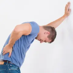 Uomo con forte mal di schiena da freddo, cerca rimedi. Si appoggia al muro con una mano, piegato dal dolore.