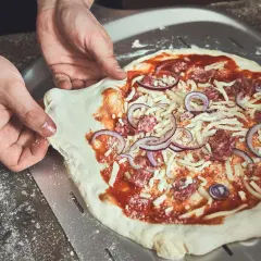 Hände ziehen den rohen Pizzateig vom Pizzaschieber. Der Teig klebt leicht am Schieber, während die Pizza mit Tomatensauce, Käse, Salami und Zwiebeln belegt ist.