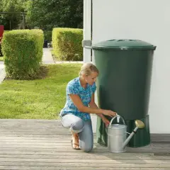 Donna raccoglie acqua piovana con un annaffiatoio da un serbatoio verde in giardino. Un modo ecologico per innaffiare le piante.