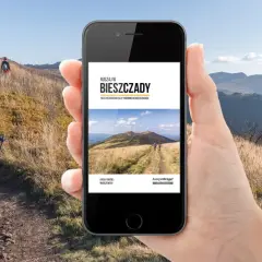Dłoń trzyma smartfon z przewodnikiem po Bieszczadach. W tle widać szlak i turystów w górach.