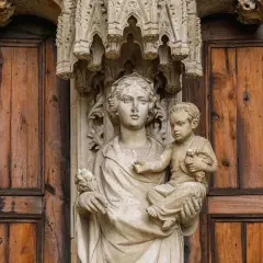 Kamienna rzeźba Matki Boskiej z Dzieciątkiem na tle drewnianych drzwi, symbolizująca teocentryzm i boską obecność w codzienności.