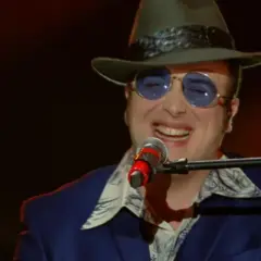 Un artista con cappello e occhiali da sole canta al microfono, un talento tra i cantanti marchigiani.