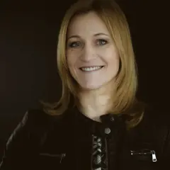 Uśmiechnięta blondynka w czarnej ramonesce na ciemnym tle.