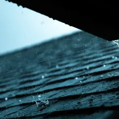 Regen prasselt auf ein dunkles Dach. Wasser tropft aus einer Rinne, ein Zeichen, dass das Blechdach reparieren nötig ist.