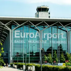 Nowoczesny budynek lotniska EuroAirport Basel Mulhouse Freiburg z charakterystyczną wieżą kontroli lotów i zielenią przed fasadą.