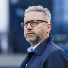 Brzęczek trener w okularach, z siwą brodą i włosami, w granatowej kurtce, patrzy w bok.