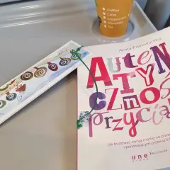 Książka "Autentyczność przyciąga" Anny Piwowarskiej na stoliku, obok ozdobna zakładka i kubek z napojami.