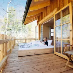 Frau liest im Bett in einer Holzhütte mit offener Tür und Blick auf Bäume.