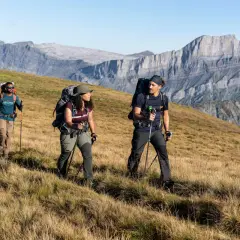 Trzech turystów z plecakami i kijkami przemierza górski szlak, przypominający **Pacific Crest Trail**.