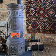 Stary, ozdobny kominek czy koza z płonącym ogniem, obok stos drewna i kolorowy kilim na ceglanej ścianie.