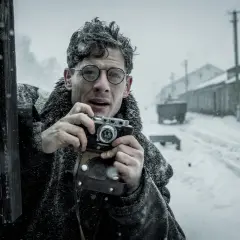 Mężczyzna w okularach z aparatem w ręku, w zimowej scenerii. Scena jak z filmów Agnieszki Holland.