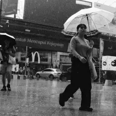 A woman with a clear umbrella walks in the rain, a tri x sign visible in the background.