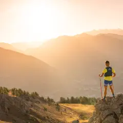 Un coureur de trail, bâtons en main, admire le paysage montagneux au lever du soleil.