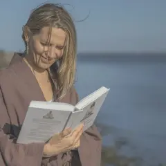 Kobieta w brązowym kimonie czyta książkę na plaży, z morzem w tle.