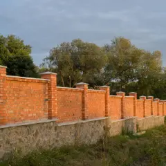 Solidne ogrodzenie murowane z cegły i kamienia, z ozdobnymi słupkami, otacza teren z zielenią i drzewami.