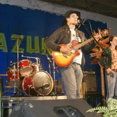 Rafał Chojnacki gra na gitarze i harmonijce ustnej, obok niego śpiewa kobieta. W tle zespół muzyczny i niebieska kurtyna z napisem "BAZUNA".