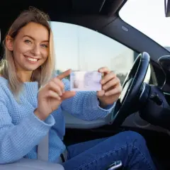 Uśmiechnięta kobieta w niebieskim swetrze pokazuje swoje nowe zdjęcie do prawa jazdy, siedząc za kierownicą samochodu.