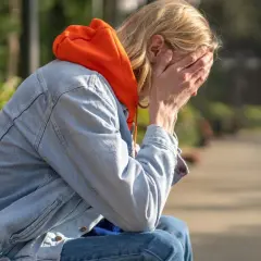 Osoba w pomarańczowej bluzie i dżinsowej kurtce siedzi na ławce, zakrywając twarz dłońmi. Może to symbolizować trudności związane z chorobą Addisona.