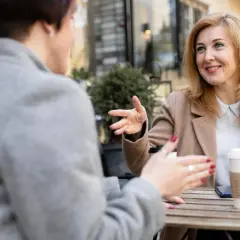 Dwie kobiety rozmawiają przy stoliku, gestykulując. Jedna z nich, z blond włosami, uśmiecha się, opowiadając o swoich przemyśleniach na temat granic w związku.