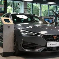 Nowoczesne konie mechaniczne, szary Cupra Leon, prezentują się w salonie. Obok stoi multimedialny kiosk.