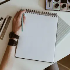 Mano con lápiz azul lista para crear dibujos de cumpleaños en un cuaderno blanco. A su lado, reglas, rotuladores y un reloj.