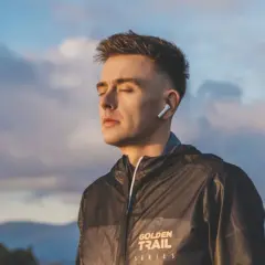 Młody mężczyzna w słuchawkach ANC, z zamkniętymi oczami, w kurtce z napisem "GOLDEN TRAIL SERIES", na tle nieba i gór.