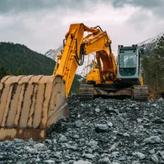 Żółta koparka na kamienistym terenie, w tle las i ośnieżone góry.