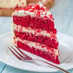 Un trozo de tarta red velvet con capas de bizcocho rojo y crema blanca, decorado con migas rojas. Un tenedor plateado espera en el plato blanco.