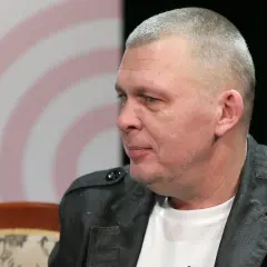 Mężczyzna z krótkimi włosami, w szarej kurtce, siedzi na kanapie. To wokalista zespołu Raz Dwa Trzy.