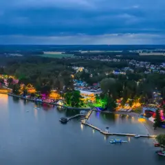Nocny widok na jezioro i rozświetlone tereny imprezowe. Widać scenę, tłumy ludzi i kolorowe światła, które tworzą atmosferę sława festiwal.