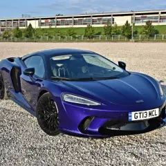 Elegancki, fioletowy McLaren GT na żwirowym placu, z nowoczesnym budynkiem w tle.