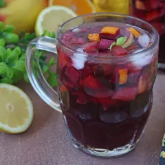 Un bicchiere colmo di sangria, con pezzi di frutta rossa, arancia e ghiaccio. La ricetta perfetta per l'estate, con limoni e foglie verdi sullo sfondo.