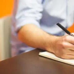 Mężczyzna pisze odręcznie notatki w zeszycie, przygotowując się do napisania prośby o dofinansowanie. Obok leży laptop.