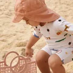 Dziecko w kapeluszu i stroju z motywem zwierząt, chroniącym przed słońcem z filtrem upf 50+, bawi się na plaży z różową łopatką i koszyczkiem.