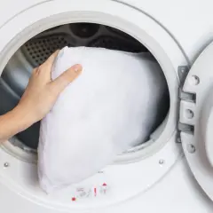 A hand places a white pillow into a washing machine, demonstrating how to wash pillows in a washing machine for a fresh, clean sleep.