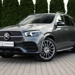 Elegancki, szary Mercedes GLE 400d SUV stoi na wybrukowanym podjeździe, otoczony gęstymi, zielonymi żywopłotami.