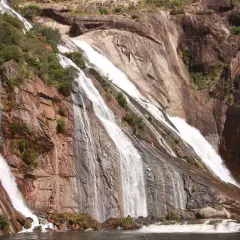 Cascada de agua blanca cayendo por rocas rojizas y grises, rodeada de vegetación. Un espectáculo natural de las fervenzas de Galicia.
