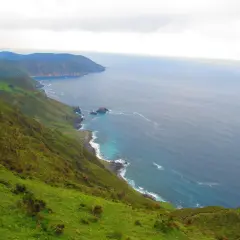 Paisaje costero verde y escarpado cerca de la garita de Herbeira, con el mar azul y las olas rompiendo en la costa rocosa.
