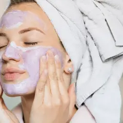 En kvinna med handduk i håret applicerar en lila ansiktsmask. En avkopplande ansiktsbehandling hemma för ren och fräsch hud.