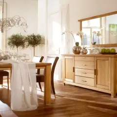 Elegancka jadalnia z drewnianym stołem i krzesłami. Nad komodą wisi duże lustro z drewnianą ramą, odbijające światło i okna.