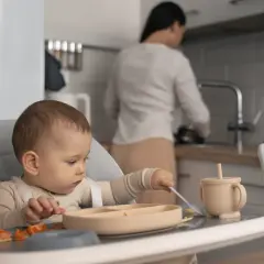 Dziecko siedzi w krzesełku do karmienia, próbując samodzielnie jeść. W tle mama przygotowuje posiłek.