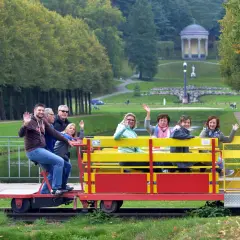 Fröhliche Menschen genießen eine Draisinenfahrt NRW durch eine Parklandschaft mit einem historischen Gebäude im Hintergrund.