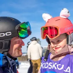 En man och ett barn i skidutrustning ler mot varandra. Barnets hjälm har söta kattöron. Perfekt för en packlista barn inför skidsemestern!