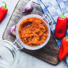 Domowy sos sriracha z papryczek chilli i czosnku w słoiku, otoczony świeżymi papryczkami i czosnkiem na desce.