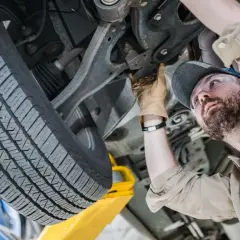 Mechanik w okularach ochronnych wykonuje sprawdzenie zawieszenia na stacji diagnostycznej. Widać jego rękę w rękawicy, która dotyka elementów podwozia.